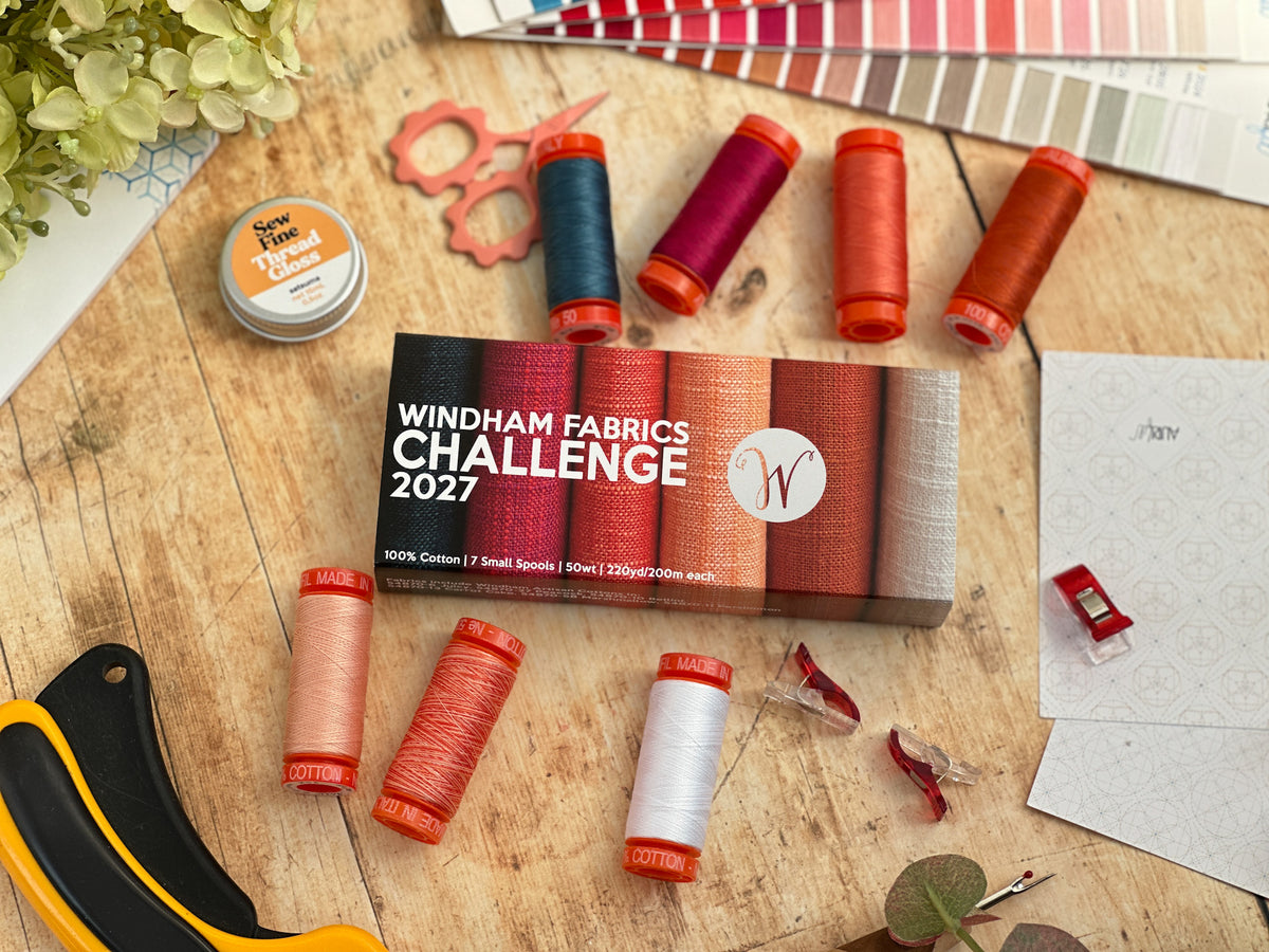 A styled flat-lay of the Windham Fabrics Challenge 2027 kit on a wooden tabletop, surrounded by thread spools in various colors, a small box of thread, a color swatch book, and crafting tools.